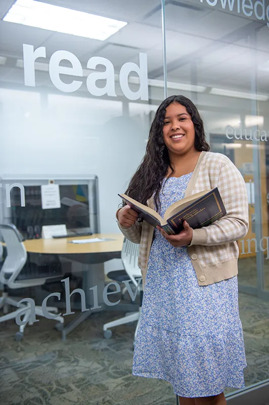A student standing in front of a glass wall with large printed words, smiling while holding an open book in a campus study area