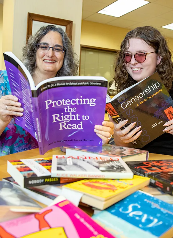 Two people sitting at a library table, smiling as they read books about censorship and the right to read, with numerous banned or challenged books spread out in front of them